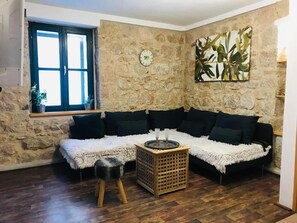 A cozy living room with a stone wall, a wooden floor, a black sofa, a white furry rug, and a wooden side table with a tray. - Stone House Saint Michael (Trilj)