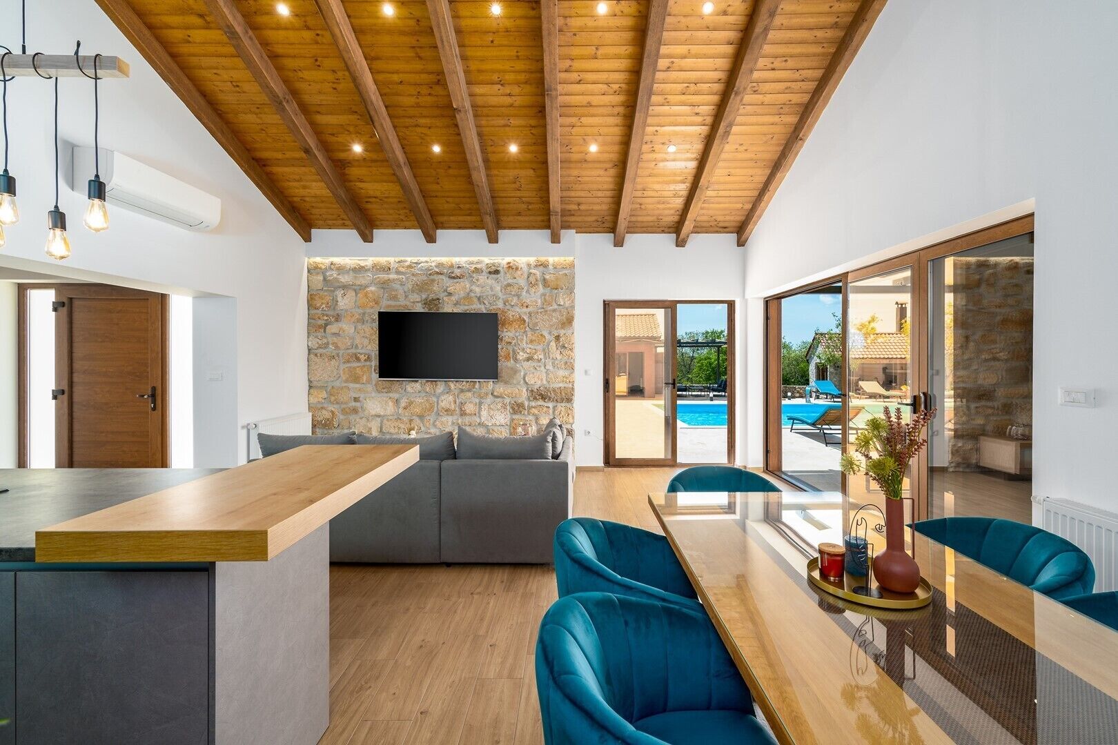 Een modern interieur met een stenen muur, houten balken in het plafond, een eethoek met blauwe stoelen en uitzicht op een zwembad.
