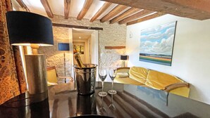 Une pièce avec un mur en pierre, des poutres en bois, une table noire avec un seau à champagne et des verres, un canapé jaune et un tableau représentant des nuages.