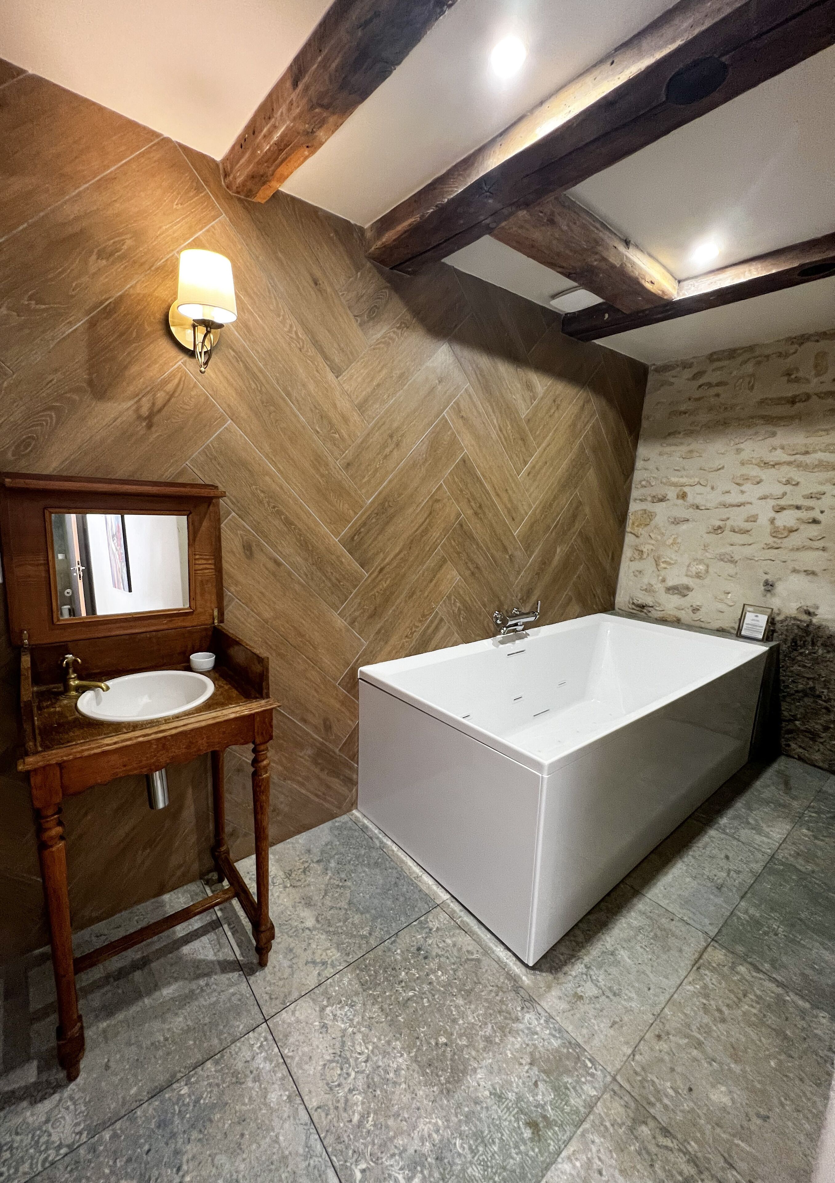 Une salle de bain avec un parquet en bois disposé en motif chevron, une baignoire blanche, un meuble-lavabo en bois avec un miroir et un luminaire fixé au mur.