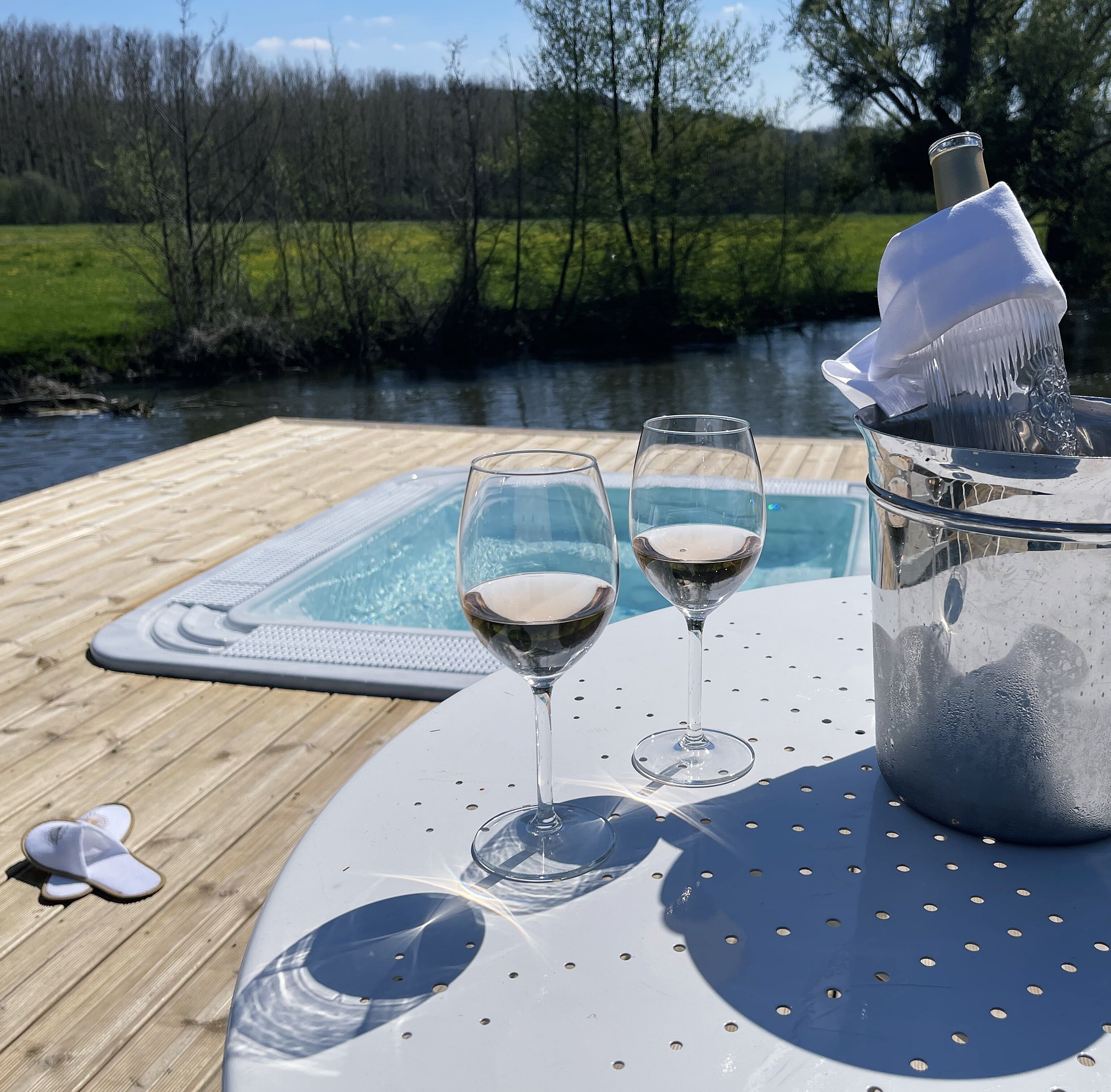 A wooden deck with a hot tub and two glasses of white wine, a bottle of champagne, and a pair of slippers.