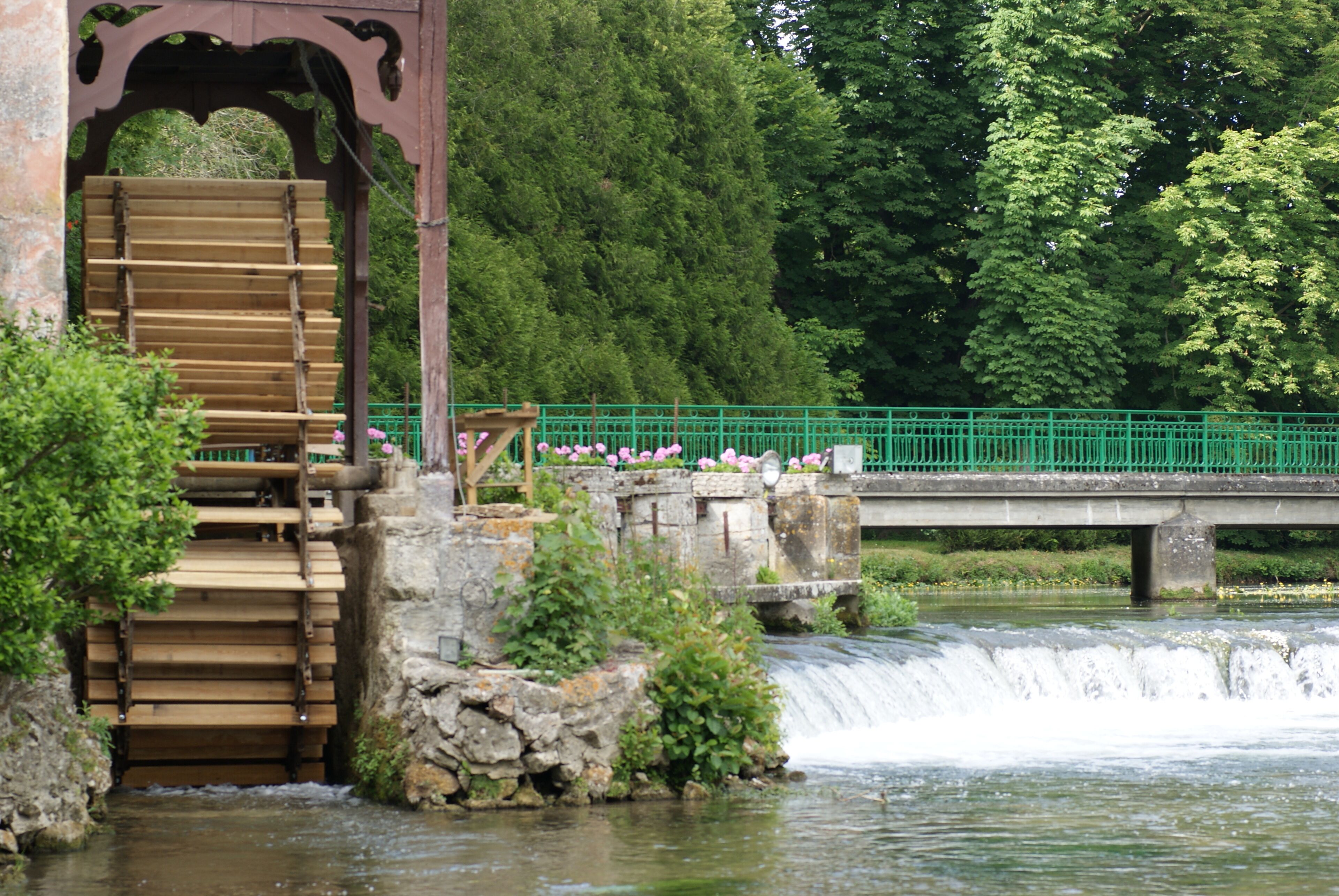 Une structure en bois abritant une roue à eau, avec des marches menant à celle-ci, un pont en pierre et une clôture verte.