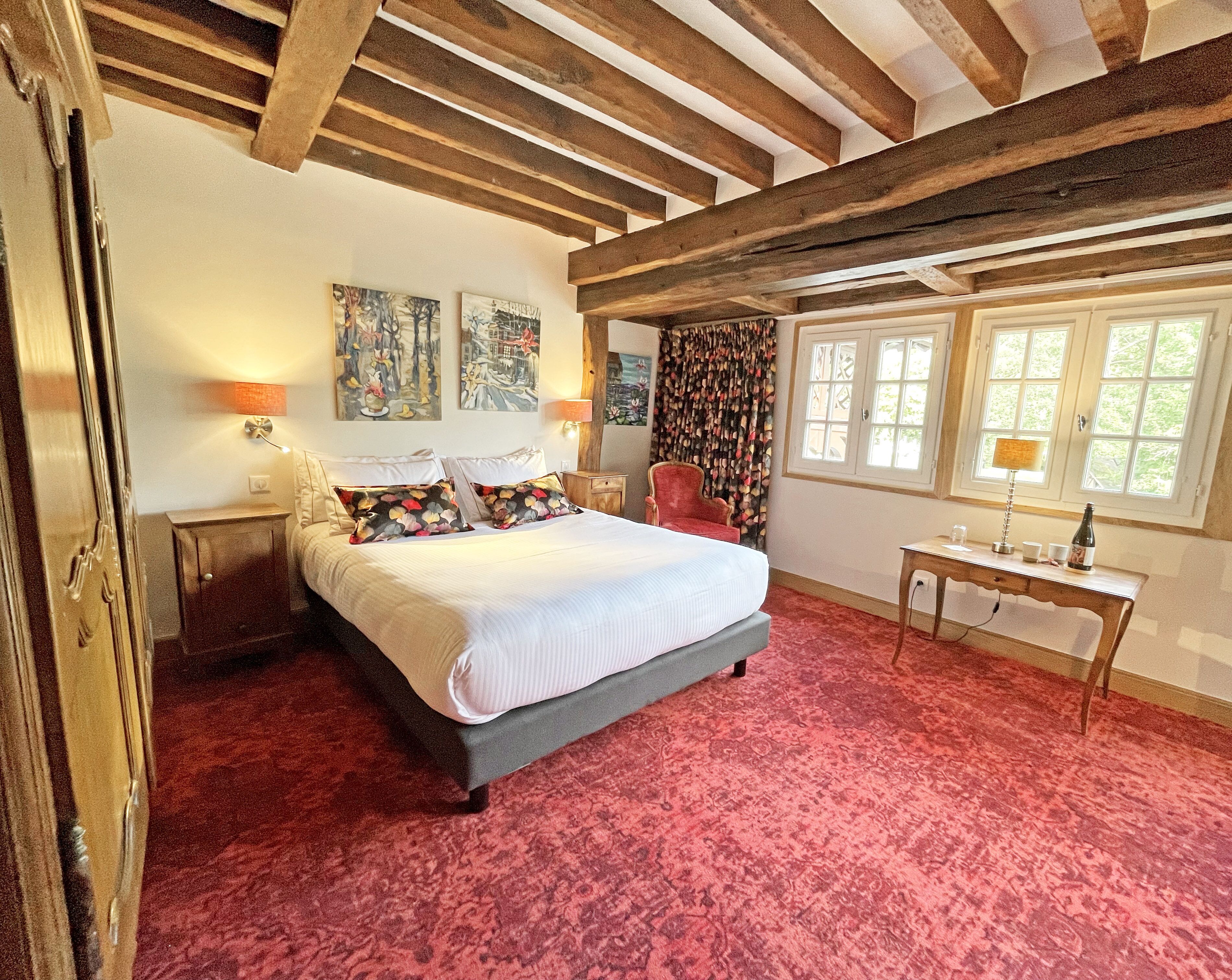 Ein Schlafzimmer mit einem großen Bett, Holzbalken, einem roten Teppich, einem Schreibtisch mit Lampe und zwei Bildern an der Wand.