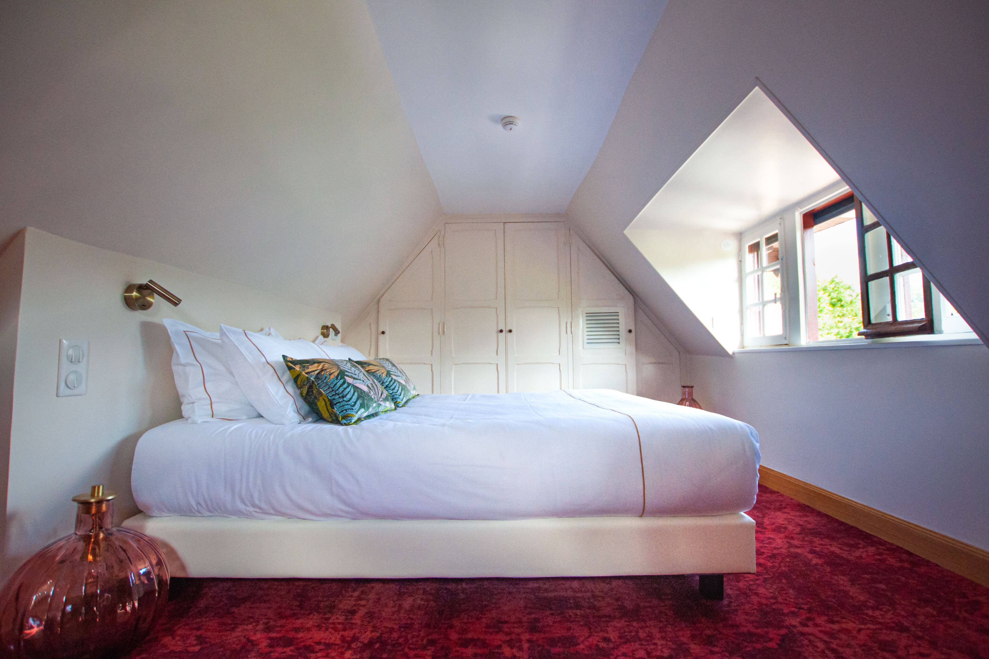 Un dormitorio con techo inclinado, una cama grande, paredes blancas y una ventana con vistas a la vegetación.