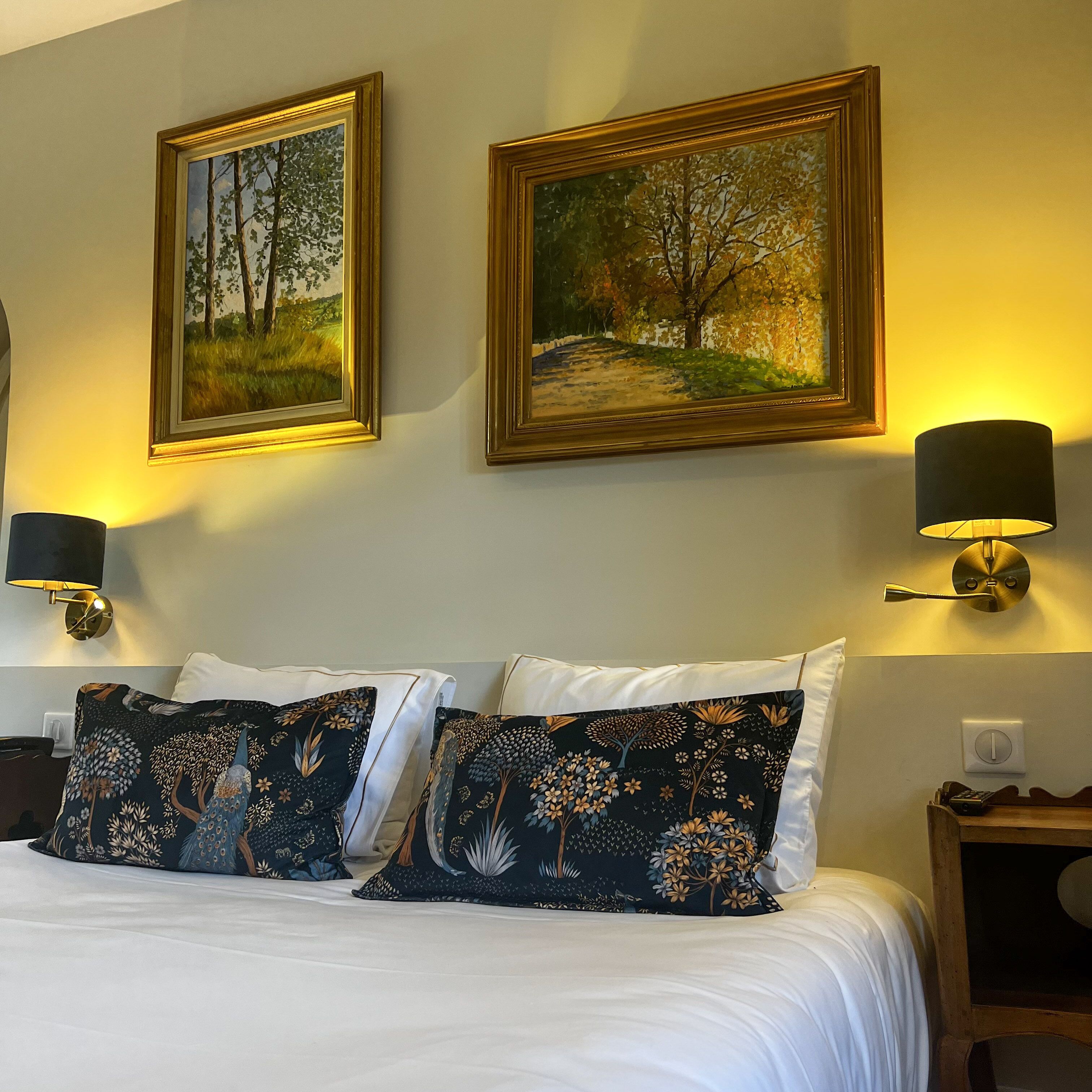 Quarto de hotel com uma cama bem arrumada, duas luminárias fixadas na parede e duas quadros emoldurados sobre a cama.