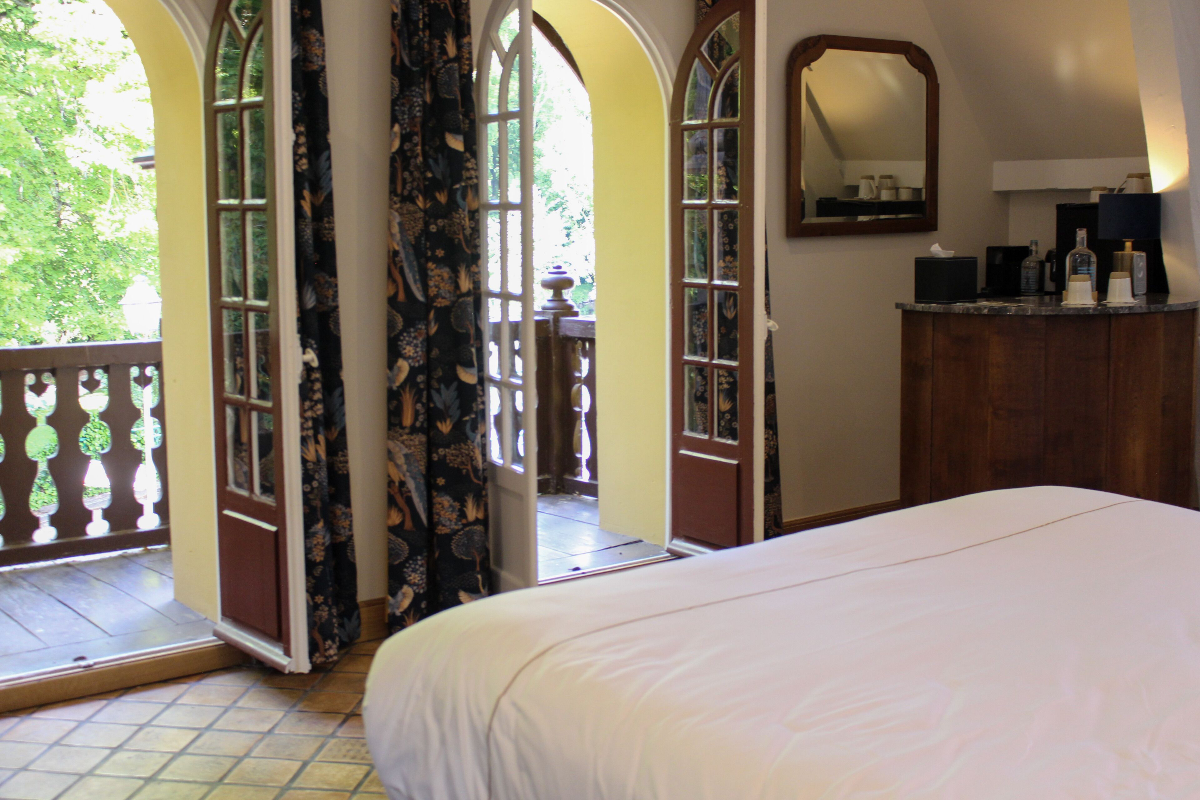 A bedroom with a large bed, a wooden dresser, a mirror, and a balcony with a view of greenery.