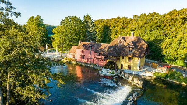 Un bâtiment historique abritant un moulin à eau, entouré d’une végétation luxuriante et bordé par une rivière.