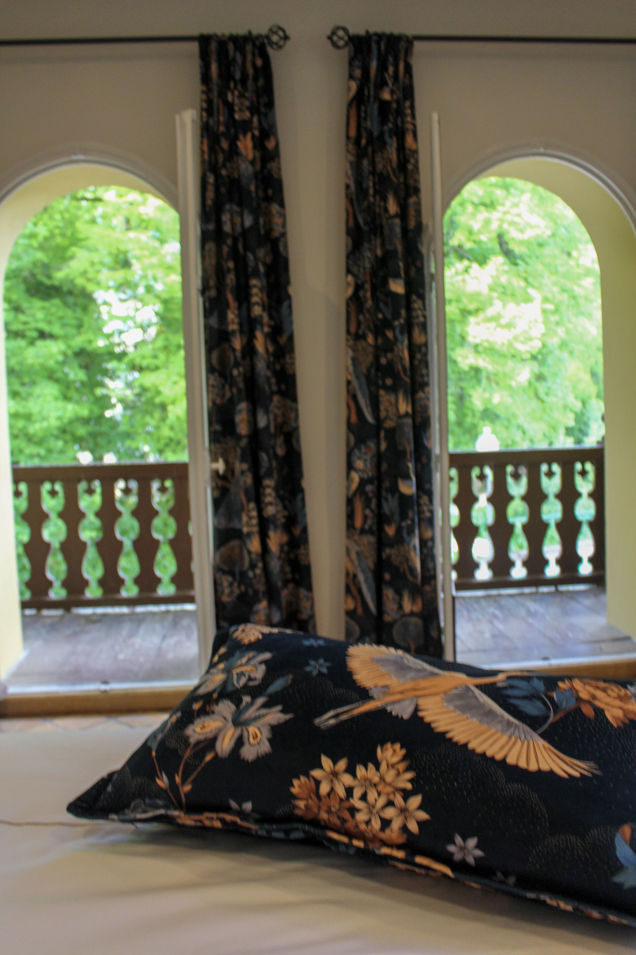Una stanza con un letto, cuscini a motivi floreali e vista sul verde attraverso finestre ad arco.