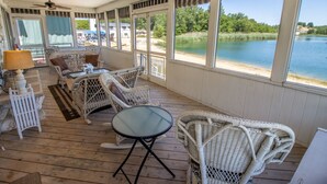 A covered porch with wicker furniture, a glass table, and large windows overlooking a lake. - 3 Bedroom | Beachwalk Resort House on Lake Kai (Michigan City)