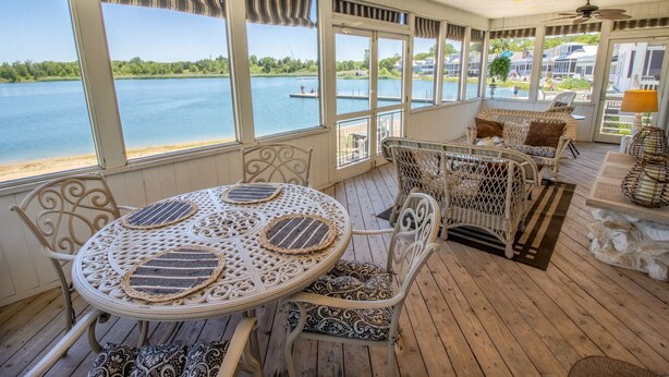 A covered outdoor seating area with a table and chairs, large windows overlooking a lake, and wicker furniture. - 3 Bedroom | Beachwalk Resort House on Lake Kai (Michigan City)
