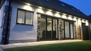 A modern house with a stone facade, large sliding glass doors, and a well-maintained lawn. - Beautiful Cosy Rural Retreat with Parking! (Winford)