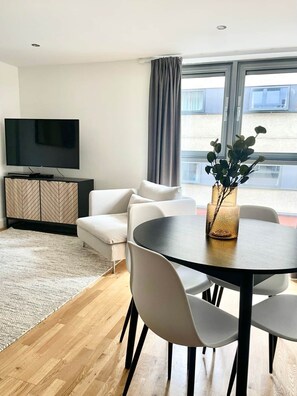 A modern living room with a flat-screen TV, a sofa, a dining table with chairs, a decorative vase, and a view of the outside through large windows.