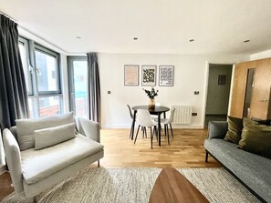 A modern living room with a grey sofa, a dining table with chairs, and a large window with curtains.