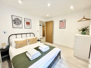 A neatly made bed with white and beige pillows, a green blanket, and folded white towels. There are framed artworks on the walls, a small bedside table with a lamp, and a potted plant on a cabinet.