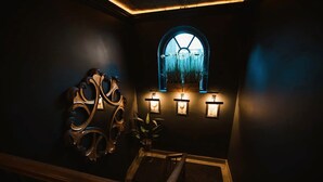 Un pasillo oscuro con una escalera de madera, un enrejado decorativo con una ventana y luces fijadas a la pared.