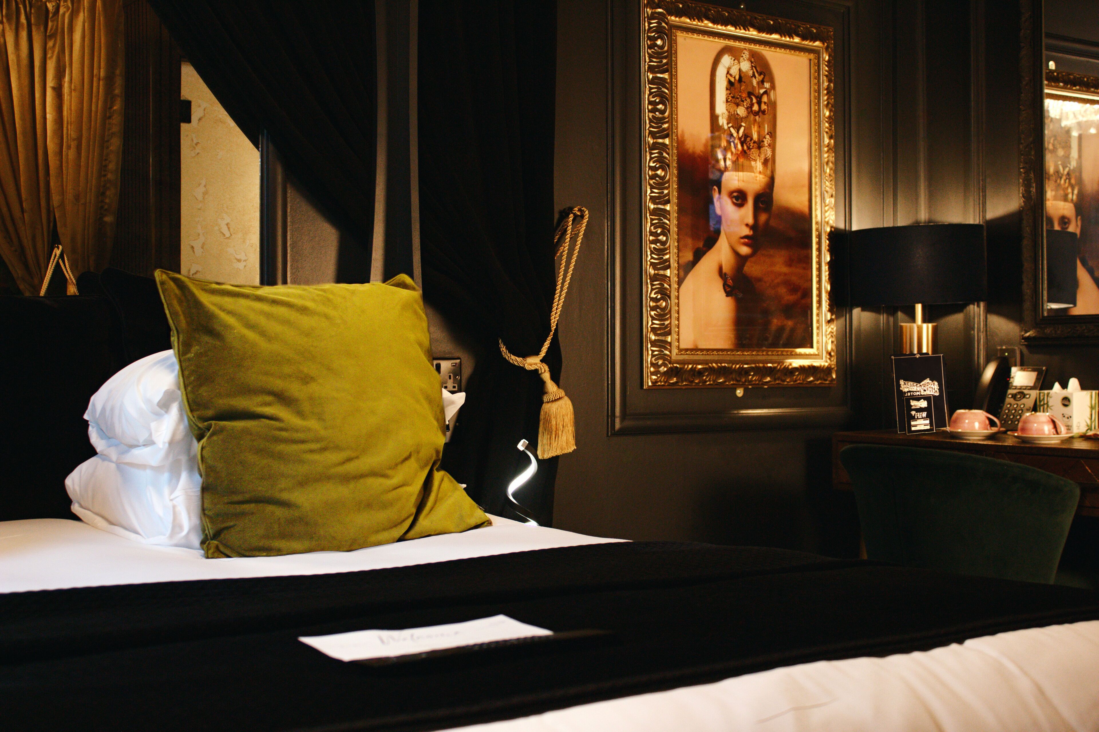 A hotel room with a bed, a green pillow, a framed picture on the wall, a lamp, and a small table with a phone and a teapot.