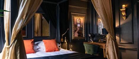 A four-poster bed with a dark blue bedspread and orange pillows, a green armchair, and a desk with a lamp.