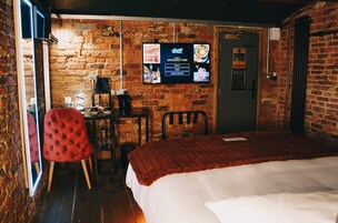 Habitación de hotel con pared de ladrillo, cama con manta roja, una silla roja, un escritorio con teléfono y televisor montado en la pared mostrando un menú.