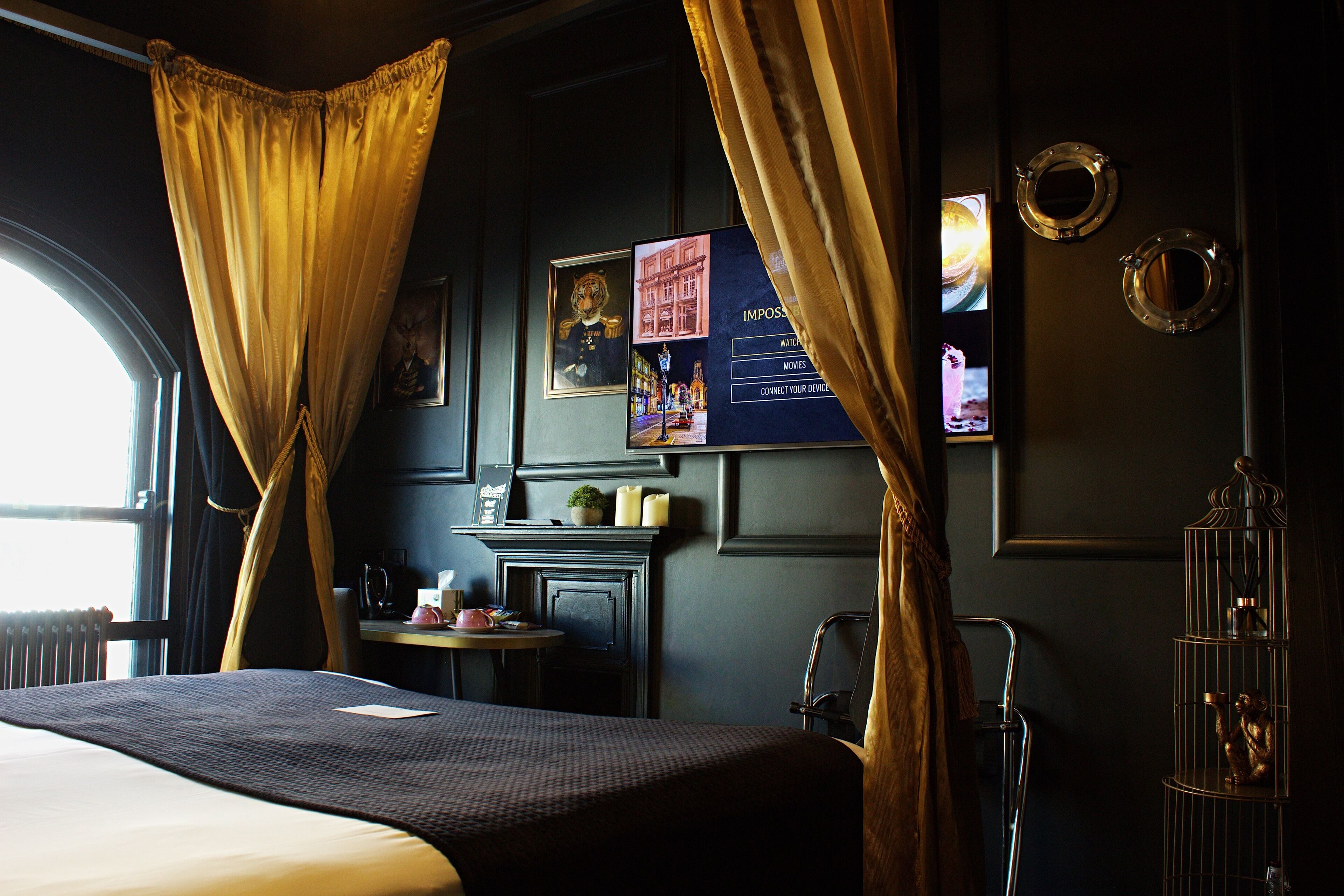 A hotel room with a bed, a small table with a lamp, a fireplace, and a large window with curtains.