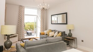 A modern living room with a grey sofa, patterned cushions, a chandelier, and a large window offering a view of greenery.