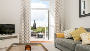 A modern living room with a sofa, a coffee table, and a TV. There is a sliding glass door leading to an outdoor area with a table and chairs.