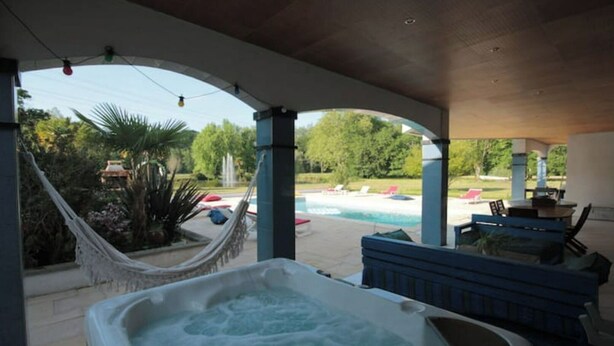 A covered outdoor area with a hot tub, lounge chairs, and a view of a swimming pool.