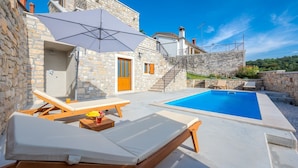 Un espace aménagé au bord de la piscine, avec des chaises longues, une table sur laquelle sont disposés des fruits et une boisson, et un bâtiment en pierre doté d’une porte en bois.