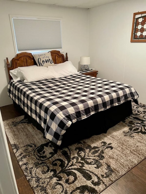 Ein ordentlich gemachtes Bett mit karierter Bettdecke, einem Holzkopfbrett, einer Nachttischlampe und einem Zierkissen mit dem Aufdruck 'let's stay home'.