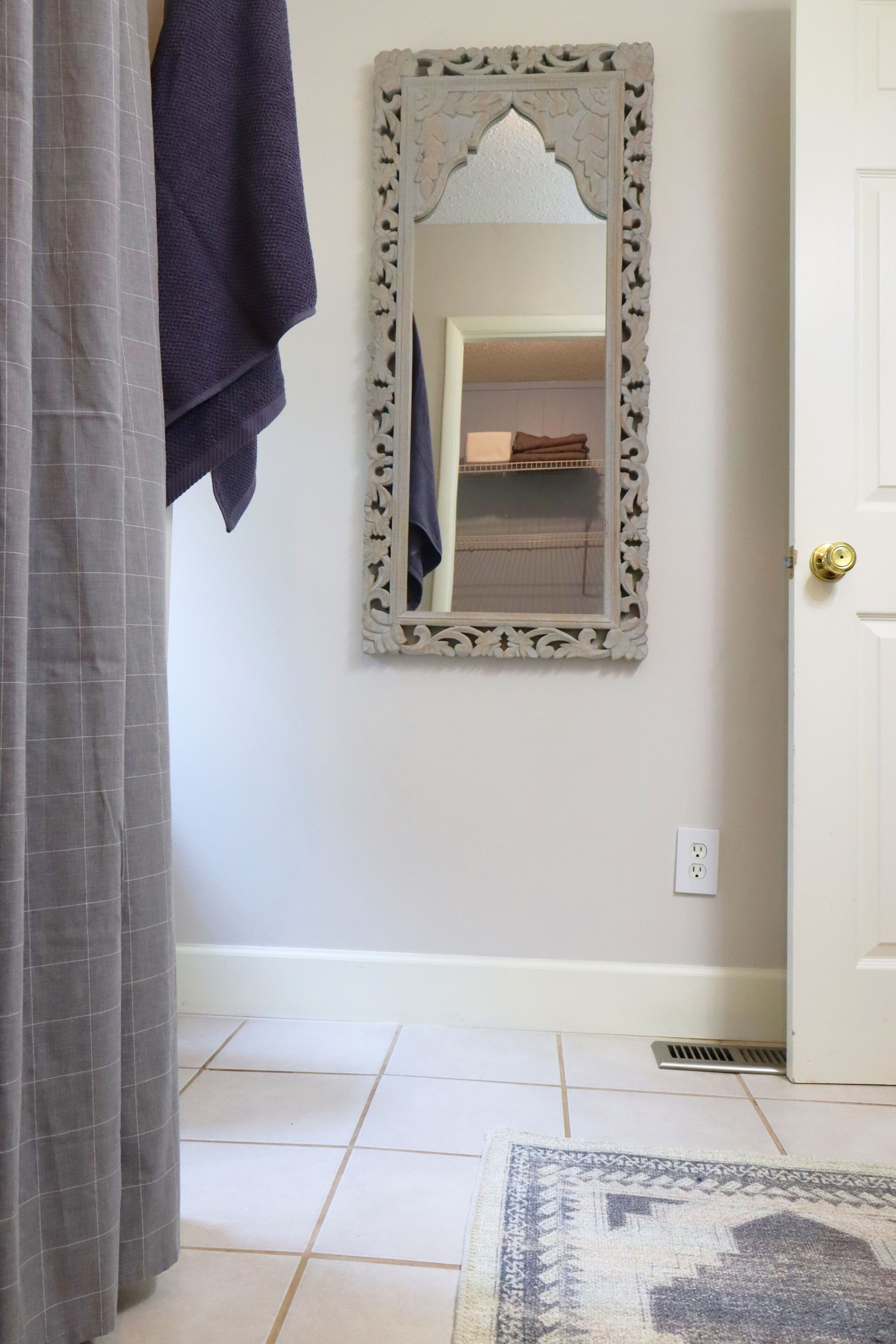 A bathroom with a white door, a mirror with an ornate frame, a towel hanging on the wall, and a patterned rug on the floor.