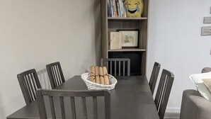 A dining table with six chairs, a basket of bread rolls, and a bookshelf in the background.