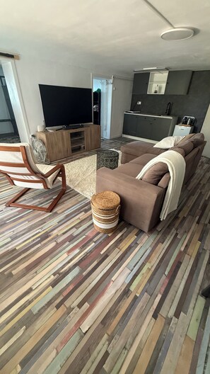 A living room with a sofa, a television, a wooden chair, and a kitchen area in the background.
