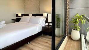 Ein modernes Hotelzimmer mit einem großen Bett, einem Nachttisch, einem am Wand montierten Kopfteil und einer Glastür, die auf einen Balkon mit Topfpflanzen führt.