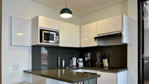 Eine moderne Küche mit weißen Schränken, einer Edelstahl-Mikrowelle, einer Kaffeemaschine und einem Mixer. Am Tresen stehen zwei weiße Hocker.