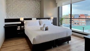 Ein modernes Hotelzimmer mit einem großen Bett, einem Nachttisch, einer Wandleuchte und Blick auf die Stadt durch große Fenster.