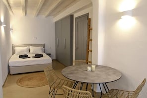 A small, well-lit room with a bed, a round table with chairs, and a door leading to another room.