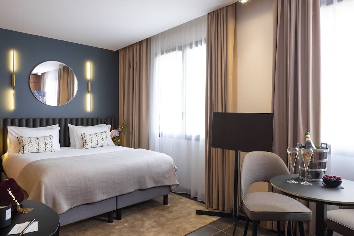 Una habitación de hotel con una cama grande, un espejo redondo, una mesita de noche con una lámpara, una pequeña mesa redonda con una silla y una ventana con cortinas.