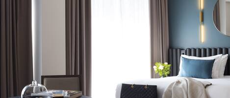 Una habitación de hotel con cama, mesita de noche con taza de café, silla y ventana con cortinas.