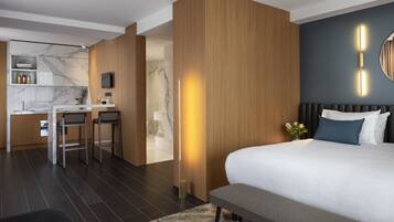 Une chambre d’hôtel moderne dotée d’un grand lit, d’un bureau avec deux chaises, d’un coin cuisine et d’une salle de bain visible à travers une porte ouverte.