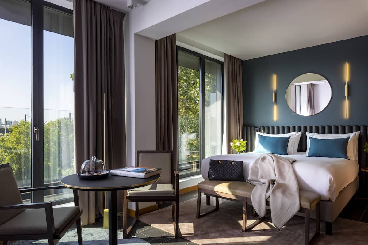 Habitación de hotel moderna con una cama grande, un espejo redondo y vistas de la ciudad a través de ventanales del suelo al techo.