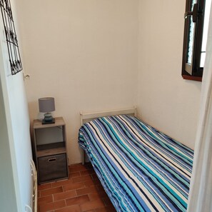 Una camera piccola con un letto singolo, una coperta a righe, un comodino con una lampada e una presa elettrica sulla parete.