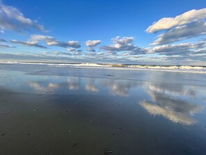 Een strand met nat zand, spiegelt wolken en golven.