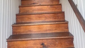 Una escalera de madera con barandilla que lleva a una puerta con una letra 'M' grande y un cartel que dice 'No se permiten zapatos'.