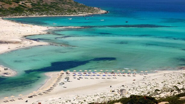 Una playa con aguas turquesas cristalinas, un acantilado rocoso y una fila de sombrillas de playa.