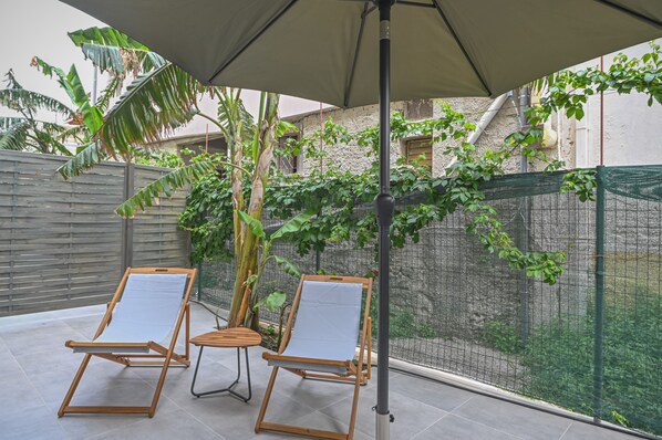 Un patio al aire libre con dos sillones de madera bajo un paraguas grande, rodeado de follaje y una cerca de alambrada.