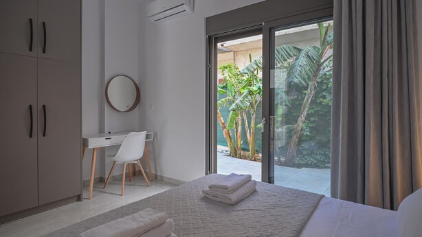 A modern bedroom with a large bed, a desk, and a chair. There is a sliding glass door leading to a balcony with plants. - Moka Apartments (Chania)
