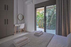 Un dormitori modern amb un llit gran, un escriptori i una cadira. Hi ha una porta corredissa de vidre que dona a un balcó amb plantes.