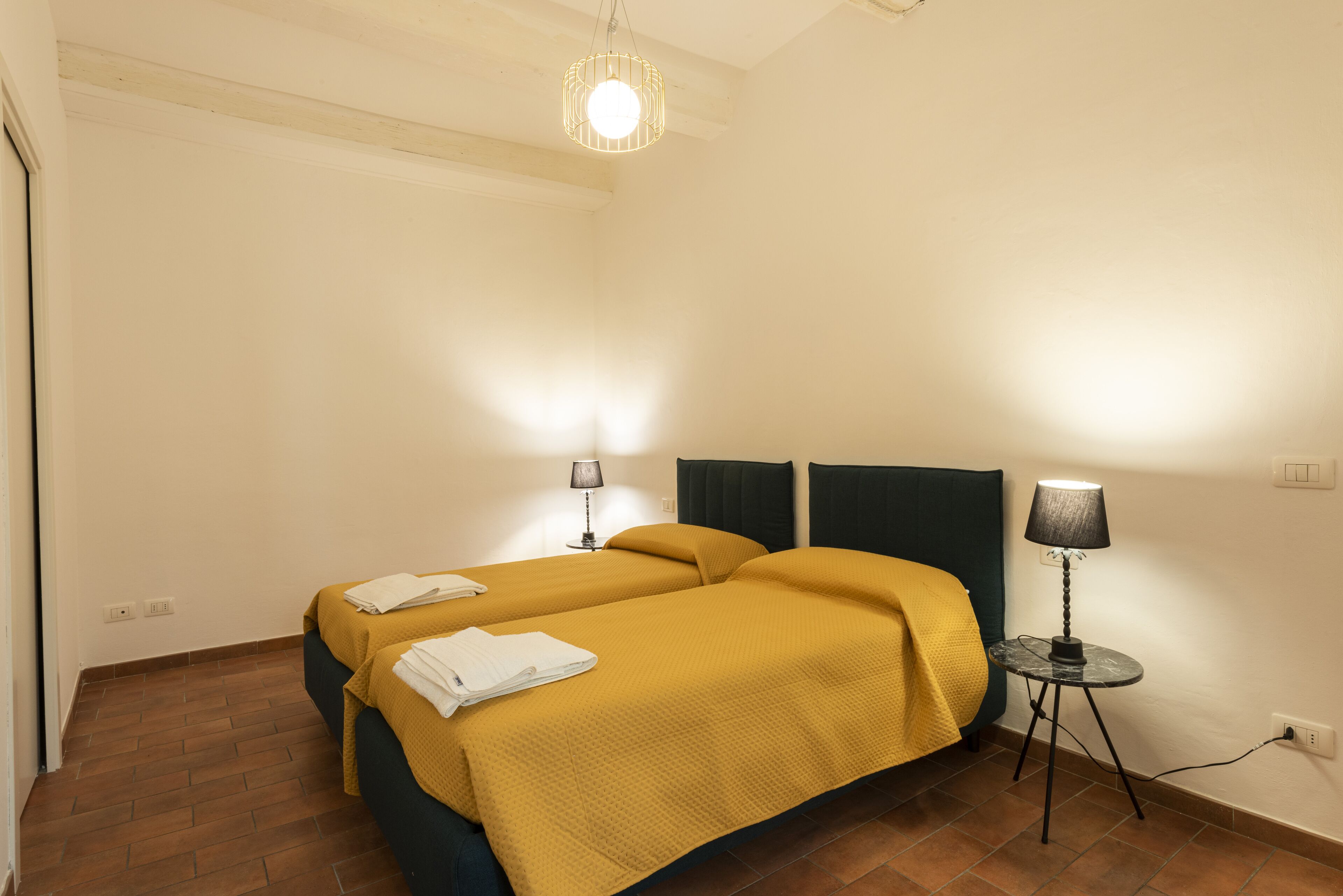 Une chambre avec deux lits, chacun recouvert d’une couvre-lit jaune et agrémenté de serviettes blanches, une petite table avec une lampe et un luminaire suspendu.