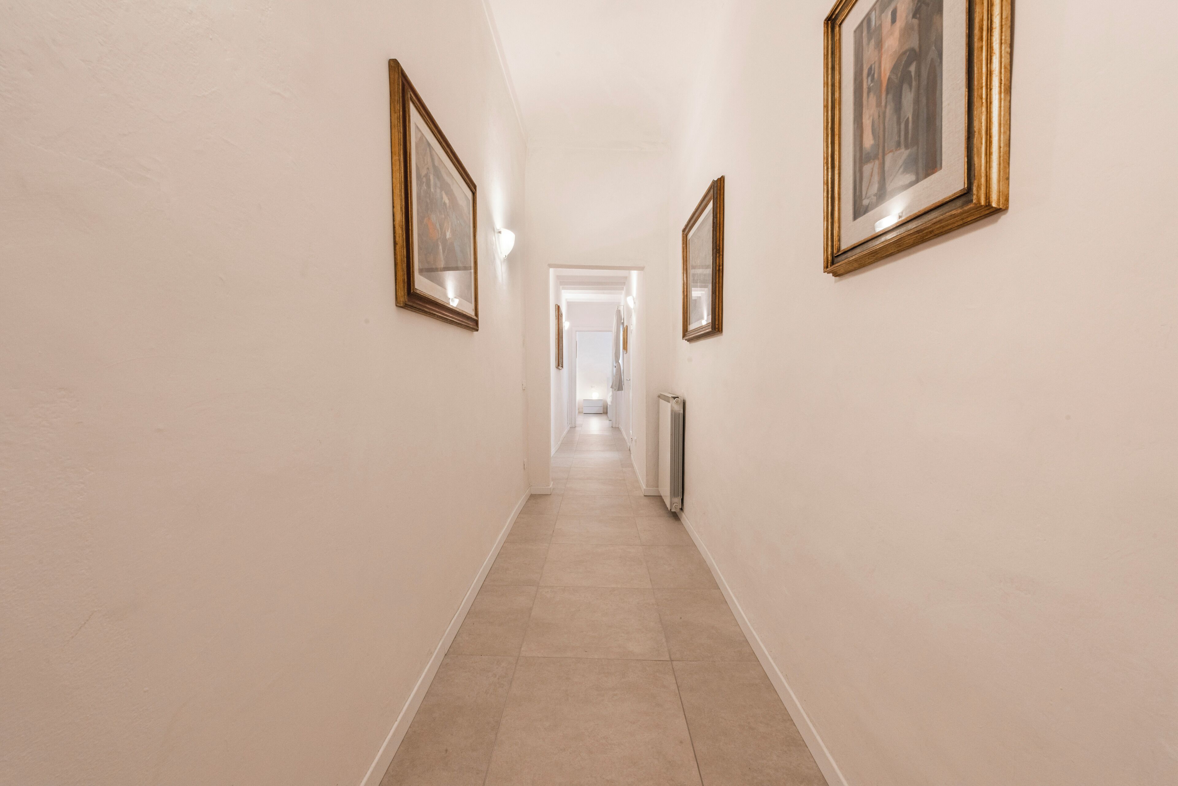 Un couloir avec des œuvres d’art encadrées accrochées aux murs, un radiateur et une porte au fond.