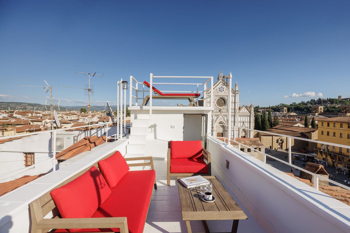 A rooftop terrace with red seating, a table with a book and glasses, and a view of a historic building with a clock tower.