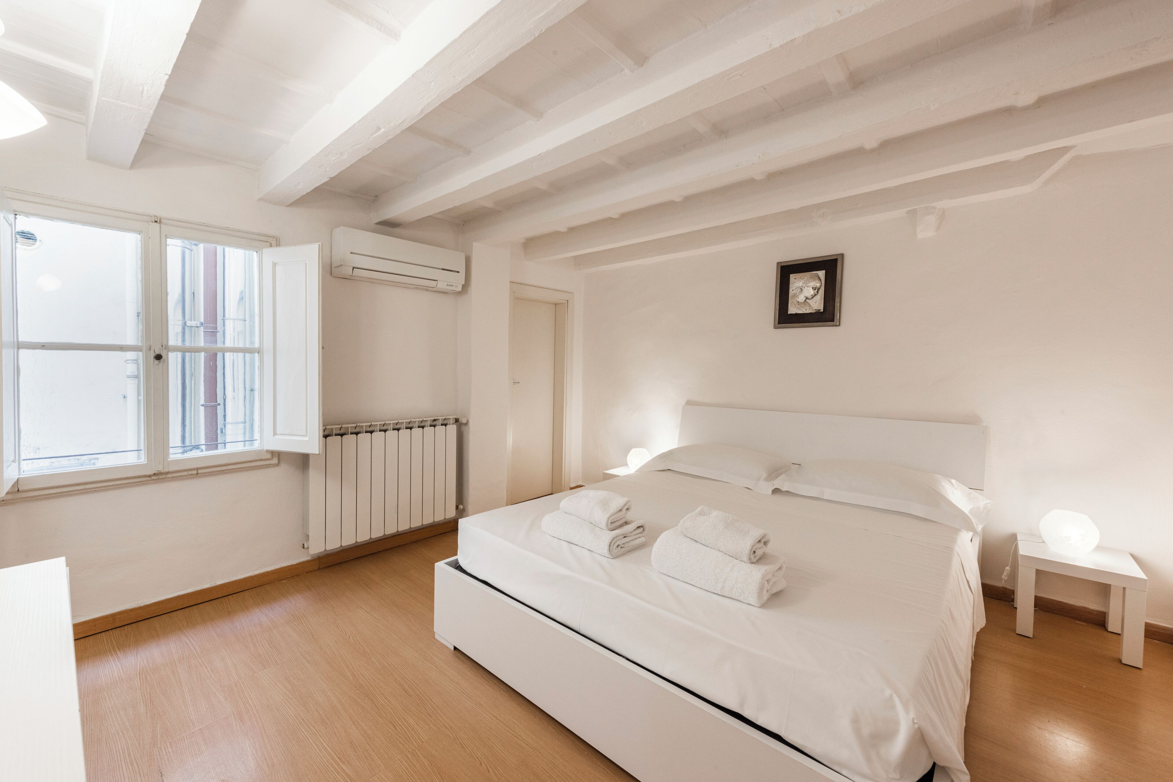 A minimalist bedroom with a large bed, a radiator, a window, and a small bedside table.